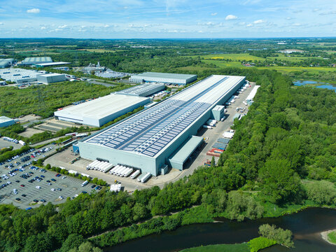 Aerial view of a large modern distribution warehouse with solar panels on the roof, white trucks at loading docks, and lush green forest bordering a river Coleshill, England, United Kingdom.