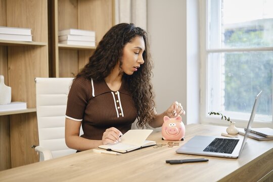 A woman sits at a wooden desk in a stylish home office, writing in a notebook while dropping coins into a pink piggy bank. Natural light filters through the window, creating a warm atmosphere