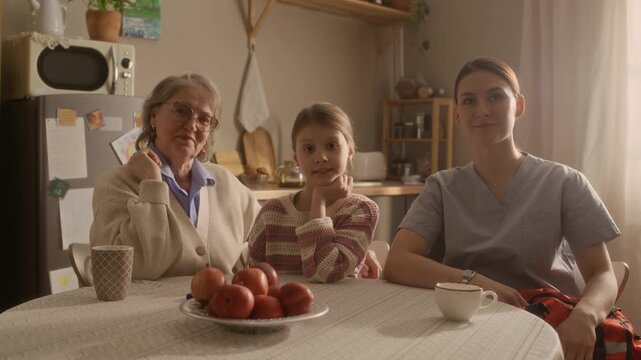 Push in shot of Caucasian grandmother, granddaughter and paramedic holding response bag while sitting at table and posing for camera