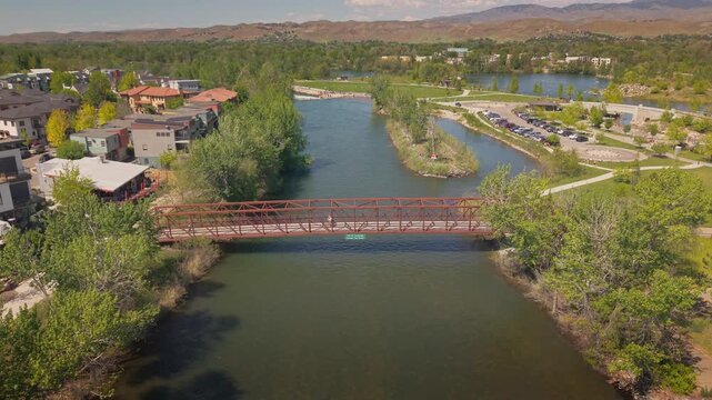 Drone flying above the Boise River toward the pedestrian bridge in Esther Simplot Park, Boise, Idaho. Bikers and walkers cross the bridge on a sunny spring day with scenic riverside views.