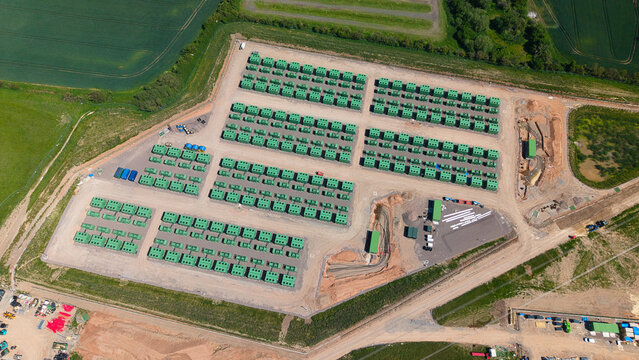 Aerial view of Battery Energy Storage Systems arranged in neat rows within a fenced industrial site surrounded by green agricultural fields in Coleshill, England, United Kingdom.