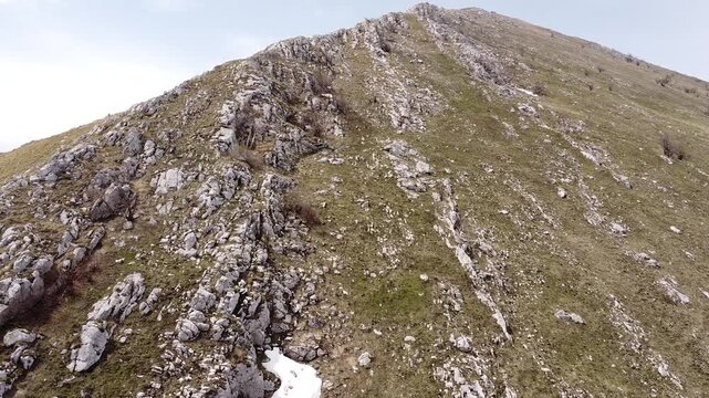 Scenic aerial footage moving over a rugged mountain ridge Rtanj, Serbia. The barren, rocky peak with patches of snow contrasts with the green slope