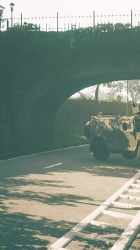 A military vehicle is parked on a road under a large bridge. The vehicle has a camouflage pattern and appears to be on patrol. Trees and grass are visible nearby.