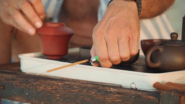 Hands arranging tea set on rustic tray closeup of skilled hands placing gaiwan and cups on weathered table by shore, casual striped shirt visible, measured movements, sunlit breeze, careful