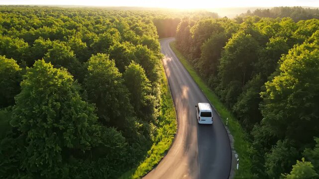 Aerial view of a white van on a winding road through a lush forest at golden hour