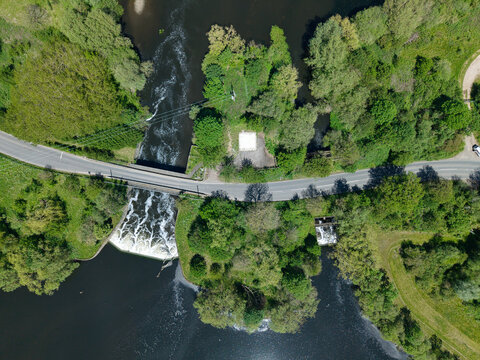 Aerial view of a river weir and a road bridge surrounded by dense green foliage and trees in Kingsbury, England, United Kingdom.
