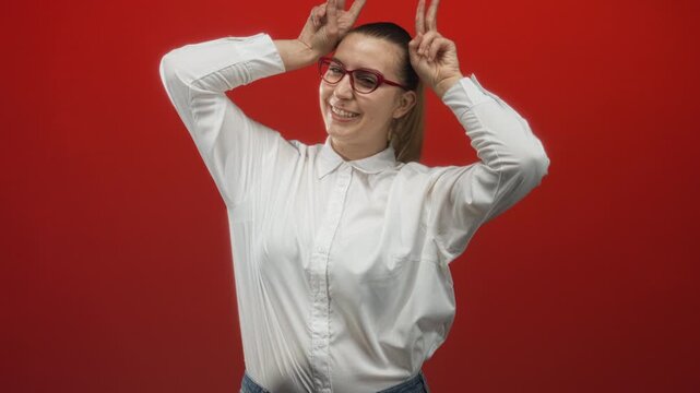 Woman wearing white shirt and red glasses with hands making bunny ears over head in studio; playfulness lighthearted charm.