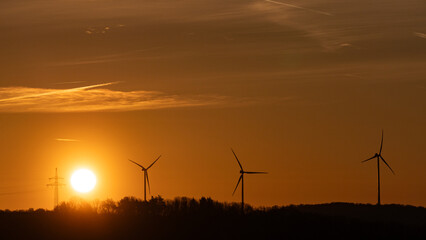 Obraz premium Wind turbine silhouettes against dramatic orange golden sunset sky with glowing sun and electricity pylon, renewable energy concept