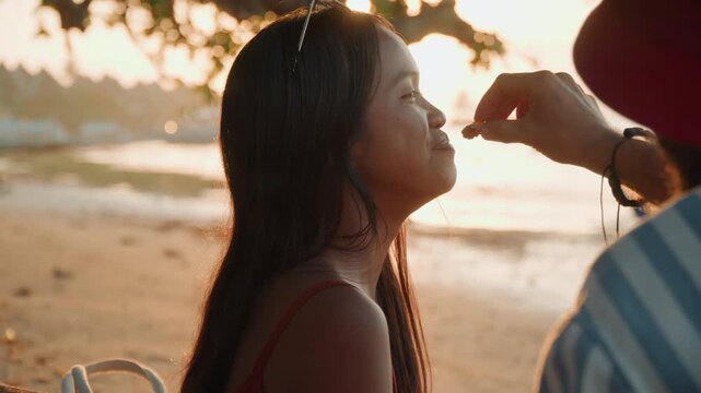 Sunset beach woman receiving makeup touch, stylist gently applies bronzer and adjusts braided hair by calm shore with boats in background, warm backlight creates intimate contemplative mood with soft