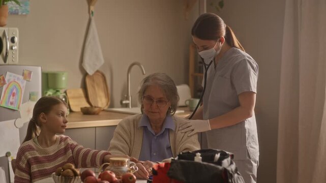 Medium shot of young paramedic performing auscultation with stethoscope, checking senior woman lungs. Little girl holding grannys hand, trying to support