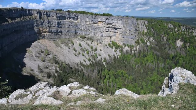Wide view of Swiss Jura cliffs and forested basin at Creux du Van in spring, with dramatic rock formations and clouds.