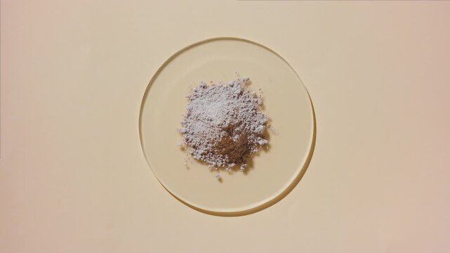 From above capture of a heap of beige fine powder on a watch glass round on a beige background lit by rotating light casting fluid, evolving shadows | Bentonite clay cosmetic ingredient concept