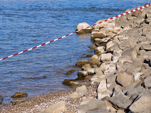 A medium horizontal shot showing a red and 
white striped warning tape running diagonally 
across a bright blue river view. The tape 
cordons off a steep bank piled with large, 
irregular grey riprap 