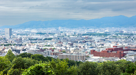 Naklejka premium Wide urban skyline of Kyoto surrounded by green hills and distant mountains during overcast daylight in Japan