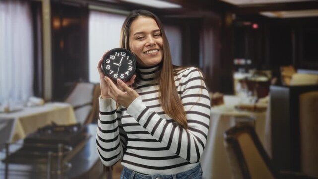 Woman holding round clock and smiling while showing hands toward camera inside dining building; time awareness joy.