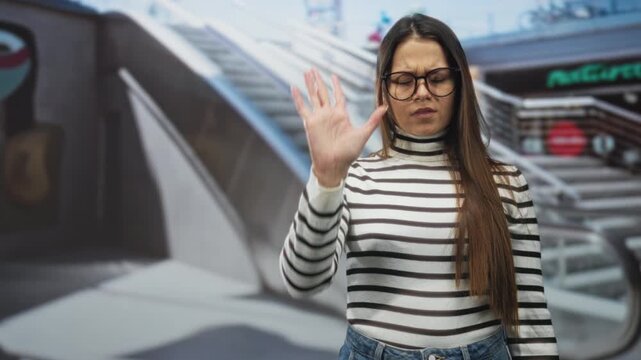 Woman in striped sweater and glasses raises her hand, palm outward and arm extended to stop in a studio with escalator mural and stairs visible; defiance boundary.