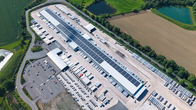 Aerial view of a large modern distribution center with solar panels on the roof and trucks at loading docks surrounded by green fields in Hinckley, England, United Kingdom.