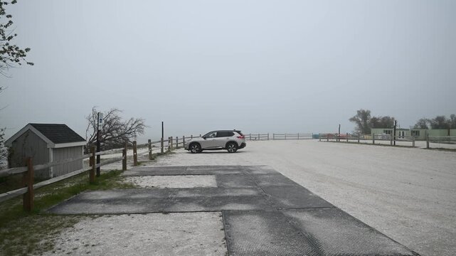 Side-view cinematic footage of an SUV slowly pulling into a fenced parking area at Assateague Island, Maryland. Dense fog blankets the nearby beach as a person dressed in black steps out of the vehicl