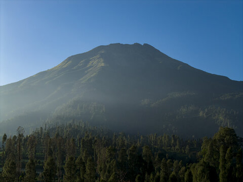 Aerial view of the majestic Mount Sumbing volcano with misty slopes and dense green forests under a clear blue sky in Kecamatan Kalikajar, Jawa Tengah, Indonesia.