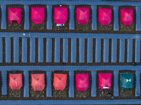 Aerial view of Garung campsite with red and orange tents arranged in rows on blue plastic-covered agricultural beds with people, Kecamatan Kalikajar, Jawa Tengah, Indonesia.