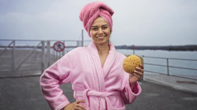Woman in pink bathrobe and towel turban holds natural sponge with hand on hip at pier, smiling at camera; self care calm.
