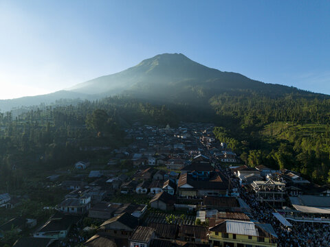 Aerial view of a large crowd gathered for Eid Al-Adha prayer in a village at the foot of a majestic mountain under a clear blue sky in Kecamatan Kalikajar, Jawa Tengah, Indonesia.