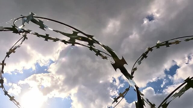 Curved barbed wire lines cross the cloudy sky while sunlight flashes between dark moving clouds. The camera shifts slowly below the fence as the wire arcs create a tense dramatic atmosphere.