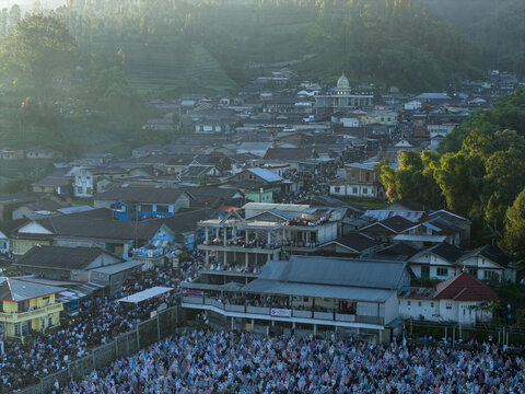 Aerial view of a large crowd of people gathered for Eid Al-Adha prayer in an open field surrounded by village houses and green hills Kecamatan Kalikajar, Jawa Tengah, Indonesia.