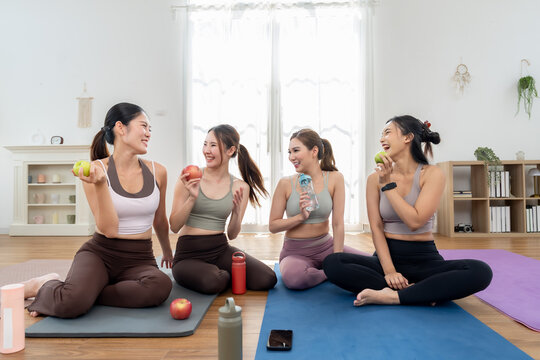 Asian female friends sit cross legged on yoga mat, laughing with apple and water bottle, enjoying post workout snack, wellness friendship, healthy lifestyle break in bright daylight studio room