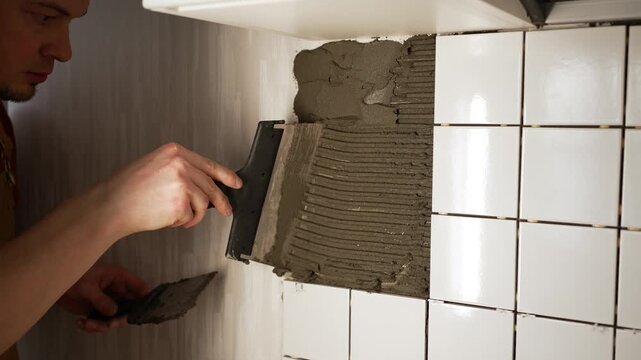 Concentrated male worker spreading tile adhesive cement mixture on the wall with a special notched trowel, preparing the surface for installing new ceramic tiles during home improvement