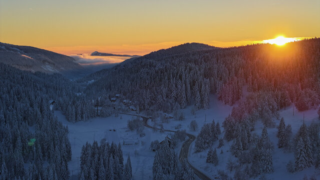 Aerial view of a snow-covered valley with dense pine forests and a winding road during a vibrant sunset in Les Rousses, Bourgogne-Franche-Comte, France.