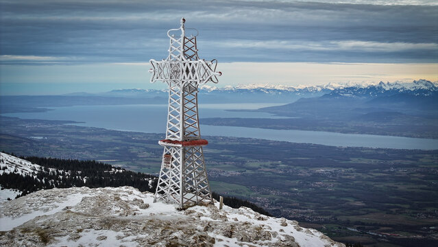 Aerial view of Le Reculet summit cross on a snow-covered mountain peak overlooking Lake Geneva and the distant Alps Thoiry, Auvergne-Rhone-Alpes, France.