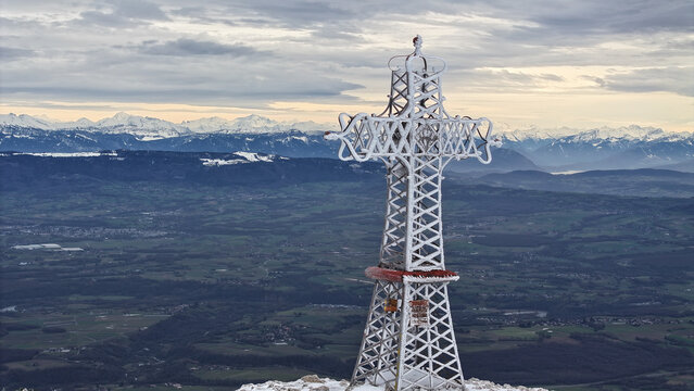 Aerial view of the frosted summit cross of Le Reculet overlooking the vast valley and the snow-capped Alps under a dramatic winter sky Thoiry, Auvergne-Rhone-Alpes, France.