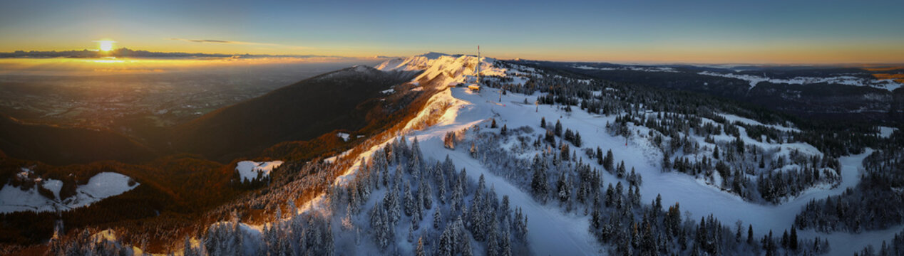 Aerial view of Col de la Faucille mountain pass with snow-covered slopes, pine trees, and a communication tower during a golden sunset Gex, Auvergne-Rhone-Alpes, France.