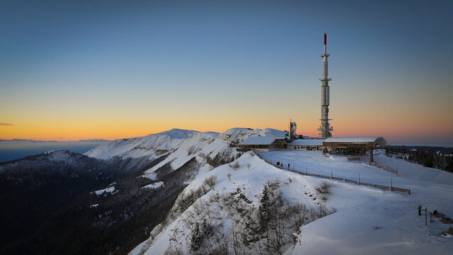 Aerial view of Col de la Faucille featuring the telecommunications tower and snow-covered mountain ridges under a sunset sky in Gex, Auvergne-Rhone-Alpes, France.
