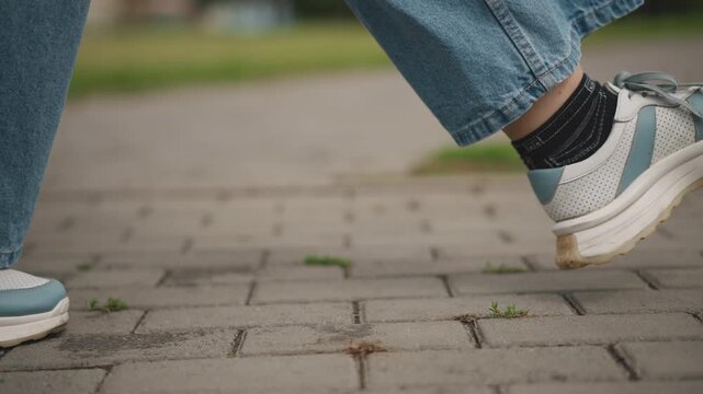 Closeup sneakers brisk stride on sidewalk, flared denim pants, confident commuting pace, urban park background, textured pavers, motion blur on toe lift, sporty streetwear energy