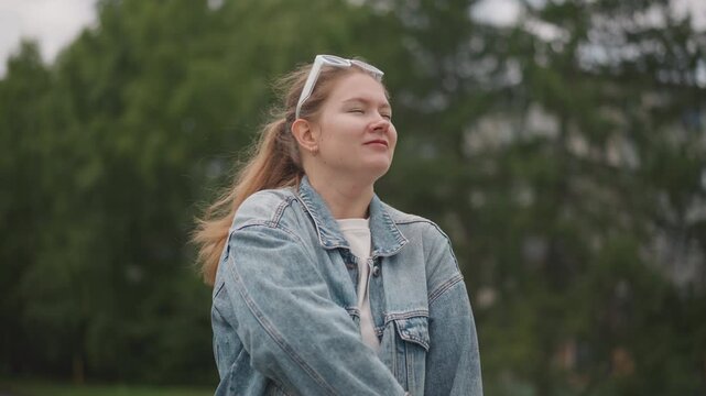 White woman in denim jacket outdoors, standing in green park carrying woven tote and adjusting strap over shoulder, calm determined gaze toward treetops, candid fashion portrait with reflective mood