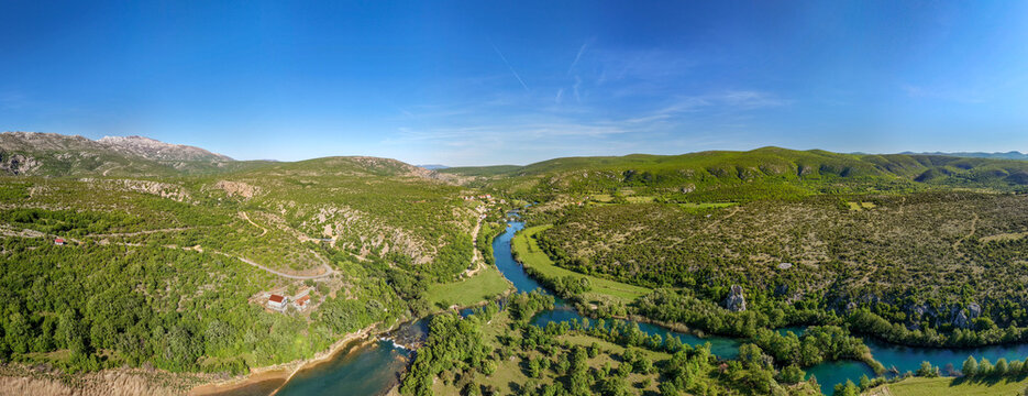 Aerial view of the Zrmanja river winding through the lush green valley of Slapovi Zrmanja with surrounding hills under a clear blue sky in Muskovci, Zadarska zupanija, Croatia.