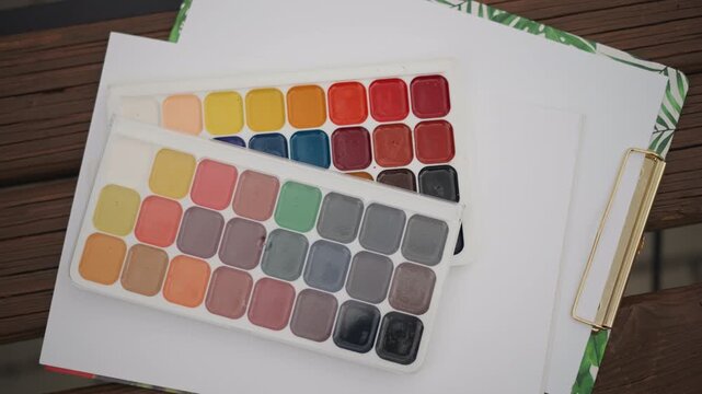 Watercolor palette on blank sketchbook page, stacked pans displaying vibrant warm and cool pigments, gold binder clip holding sheets, wooden tabletop backdrop, overhead flatlay with soft natural