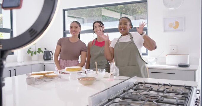 Diverse female friends in aprons greeting ringlight phone filming presenting cake on kitchen island