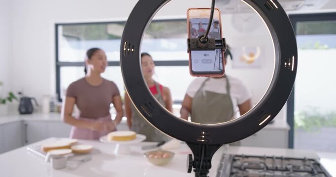 Diverse female friends assembling cake at kitchen island, smartphone on ring light filming tutorial