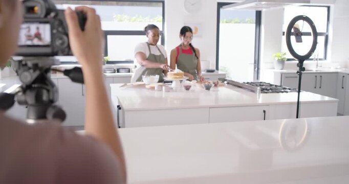 Diverse female hosts in aprons icing cake at island while operator framing DSLR ringlight filming