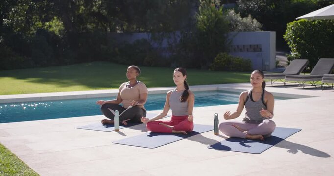 Diverse female friends following guide, flowing into meditation mudra on mats with bottles poolside