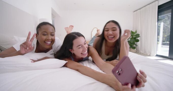 Diverse female friends in bedroom center woman reaching and taking selfie on phone capturing moment