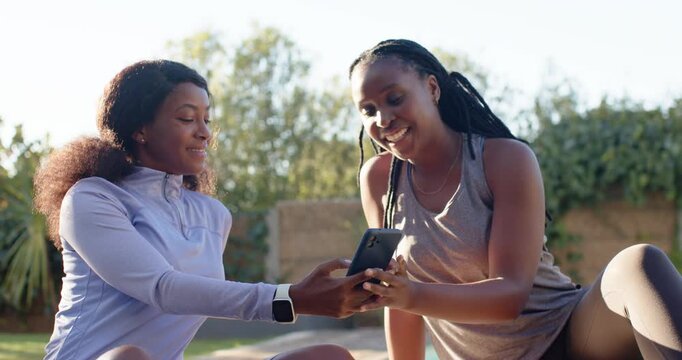Two adult women sitting on poolside mat in activewear offering smartphone, reaching to view content