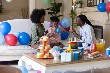 Obraz premium African American family sitting on white sofa celebrating birthday with cake, balloons, gifts