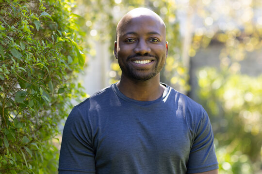 African adult man smiling and standing in sunlit backyard wearing navy shirt near hedge