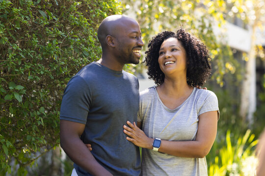 African American couple laughing and standing along neat hedge in sunlit garden, showing smartwatch