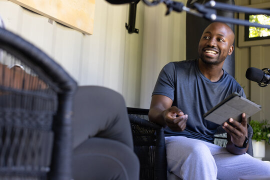 African American man sitting in pod nook wicker seat gesturing holding tablet talking into boom mic
