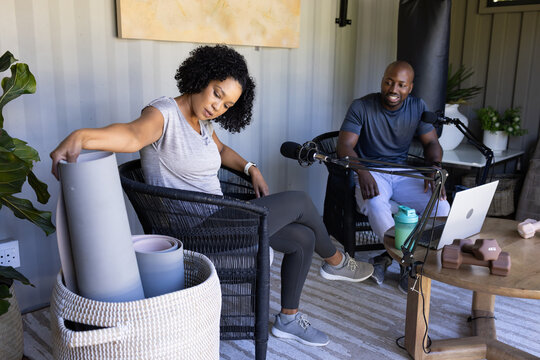 African American couple in gymwear sitting in home studio, woman reaching into basket, mics laptop